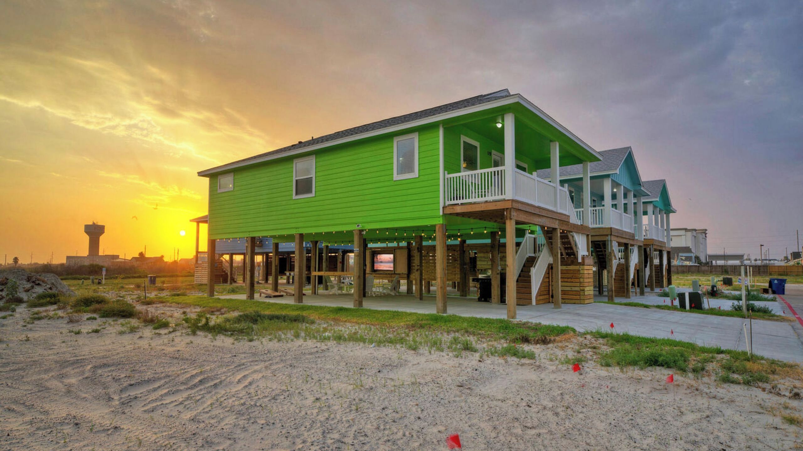 A green two-story house with a balcony, wooden deck, and a view of the sunset.