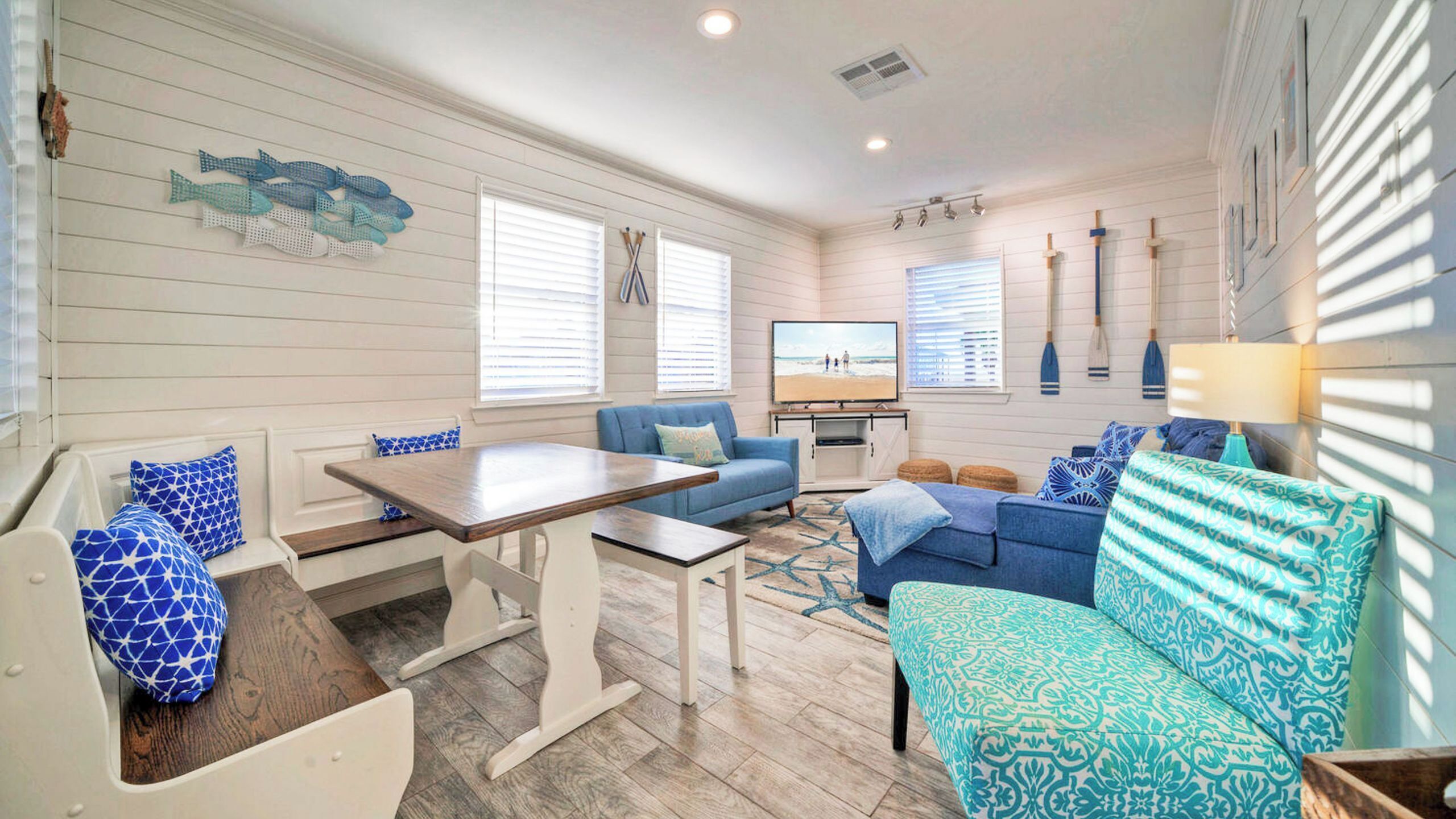 A room with a wooden bench, a blue sofa, a patterned ottoman, a dining table, and a TV.