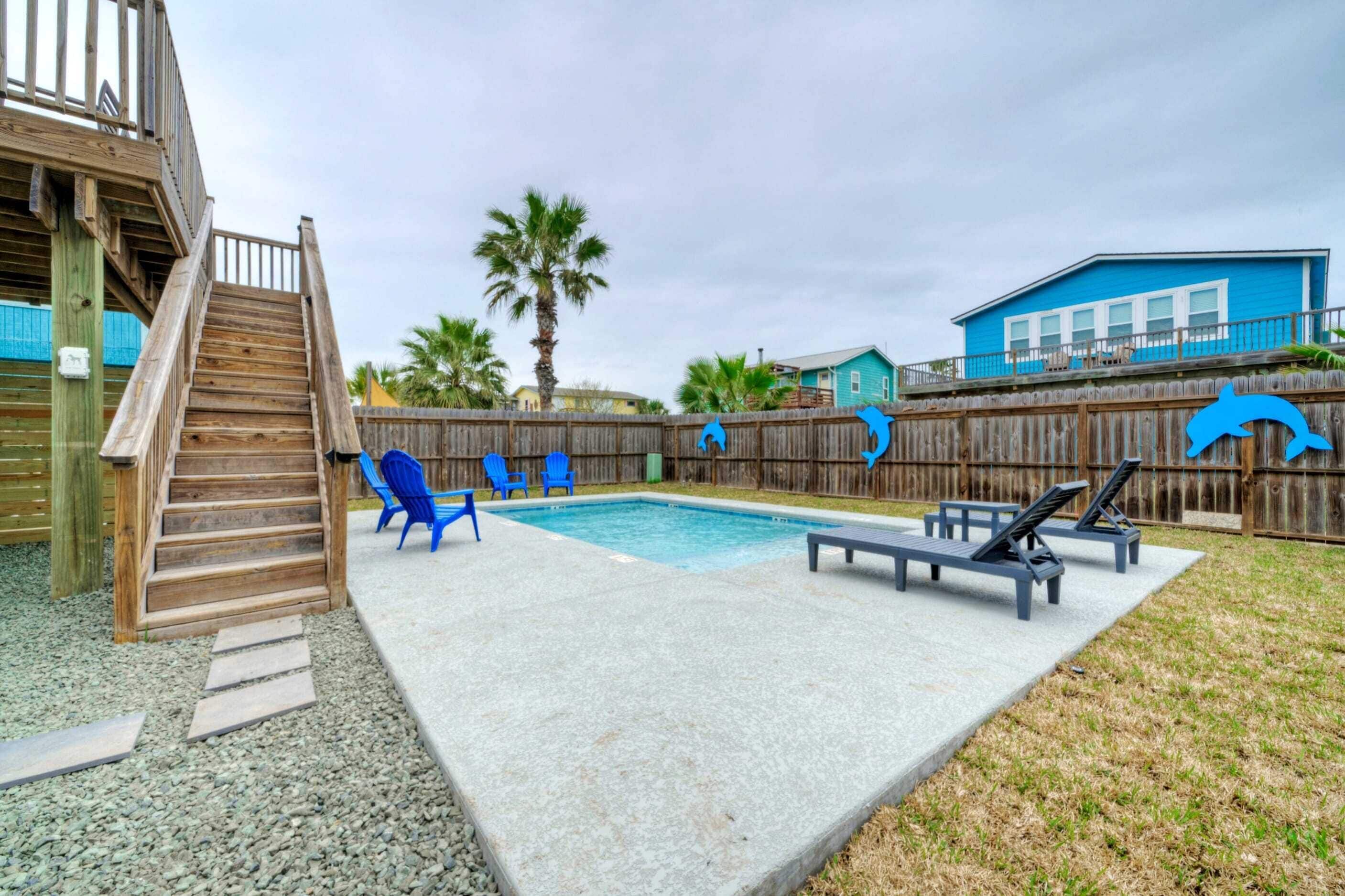 A backyard with a swimming pool, a wooden deck, and a fenced area with blue chairs.