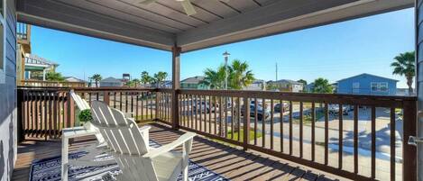 A covered outdoor area with a wooden deck, a ceiling fan, and a view of a parking lot and houses in the background.