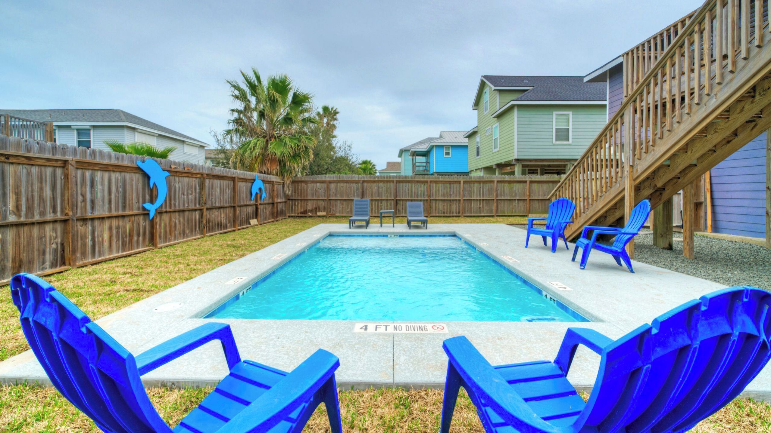 Una piscina rectangular con muelle de madera, sillas azules y una cerca de madera.