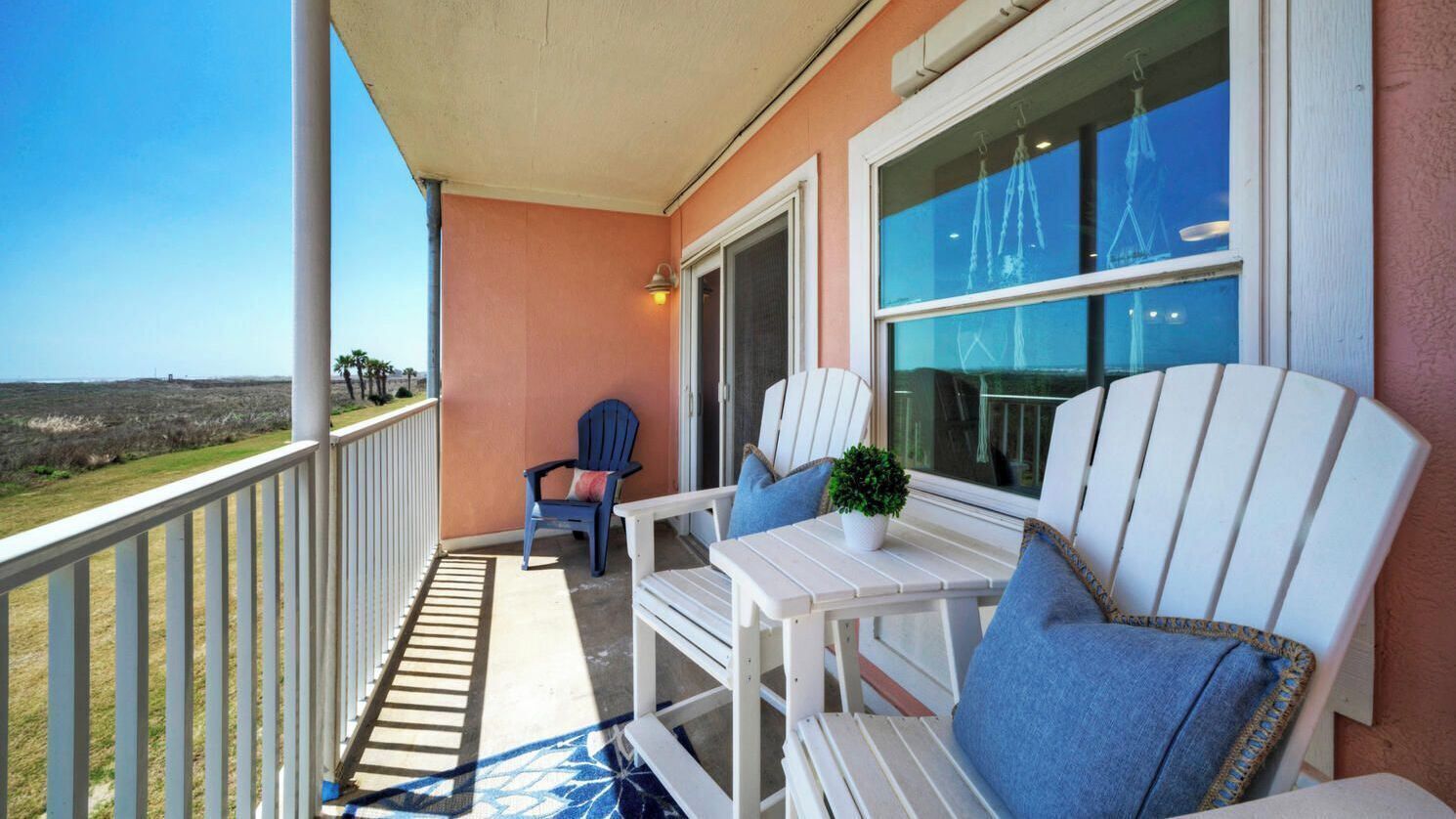Een balkon met een witte railing, een blauwe stoel, een witte tafel met een plant, en uitzicht op het landschap.