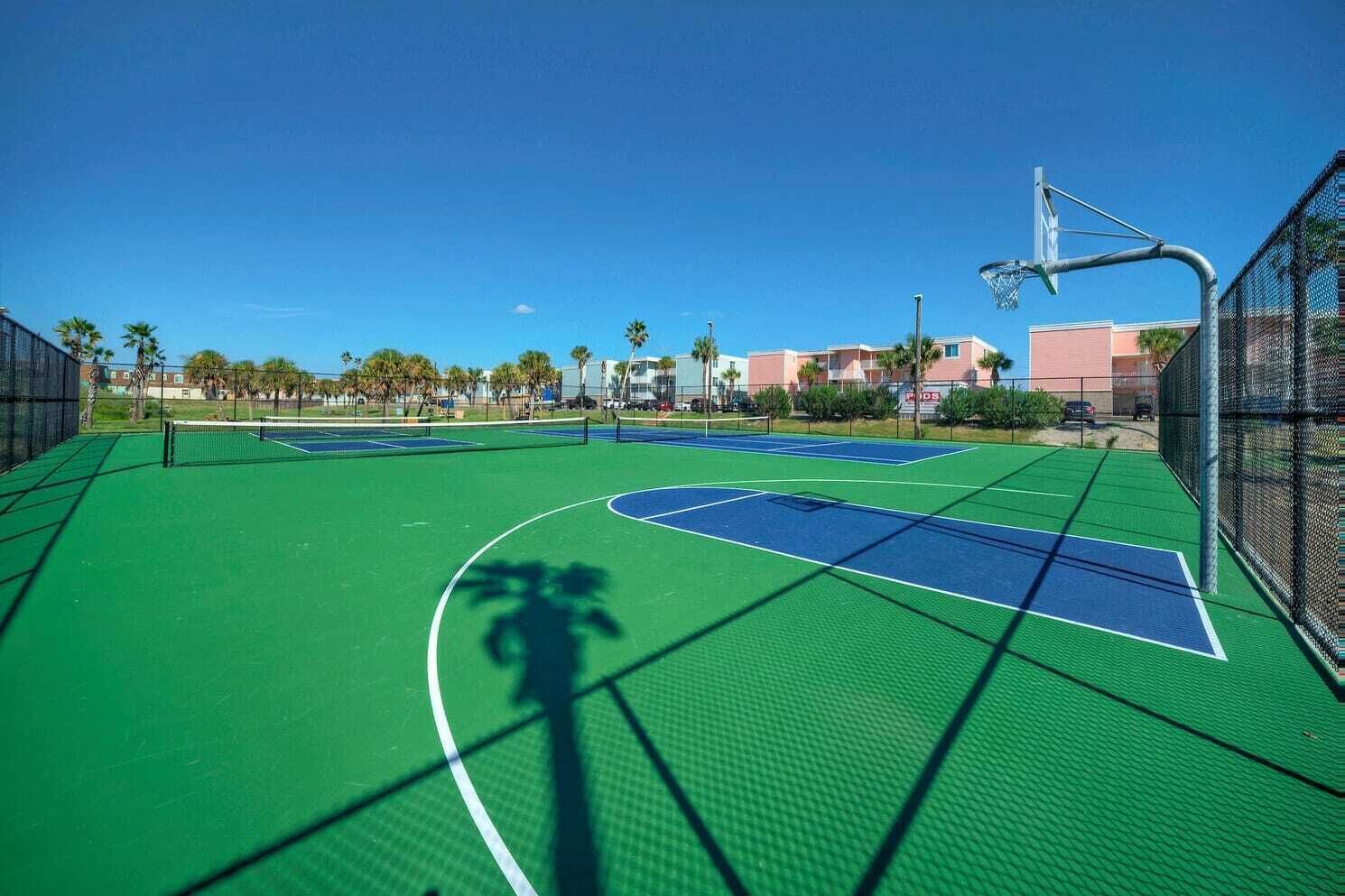 An outdoor tennis court with a net, surrounded by a fence and palm trees.
