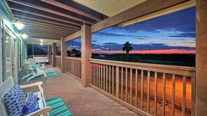 A covered outdoor area with wooden deck, white chairs, and a view of the sunset over the ocean. - Incredible Nature Views, Golf Cart for Rent, Balcony Blue Crush by Avantstay (Port Aransas)