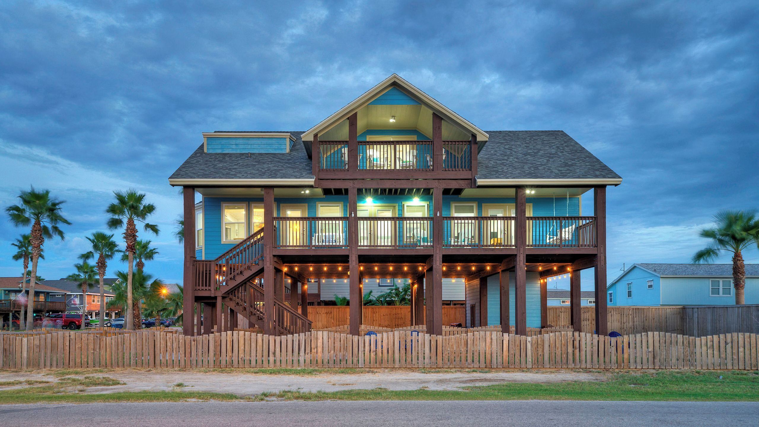 A two-story beachfront house with a balcony, wooden deck, and a fenced front yard.