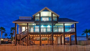 A two-story beachfront house with a wooden deck, multiple balconies, and string lights.