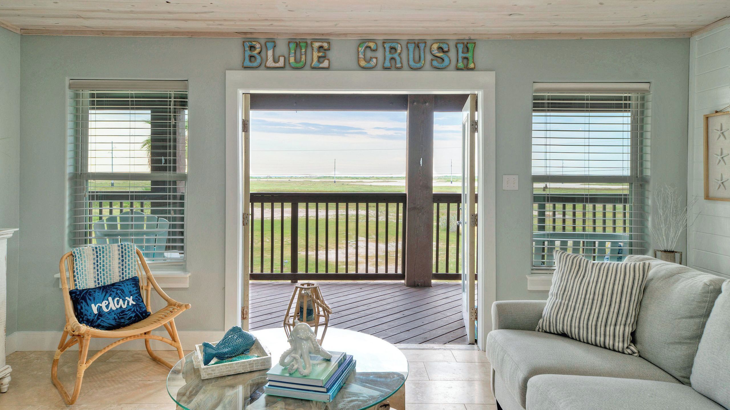 A room with a view of the beach, a wicker chair, a glass table with decorative items, and a sofa.