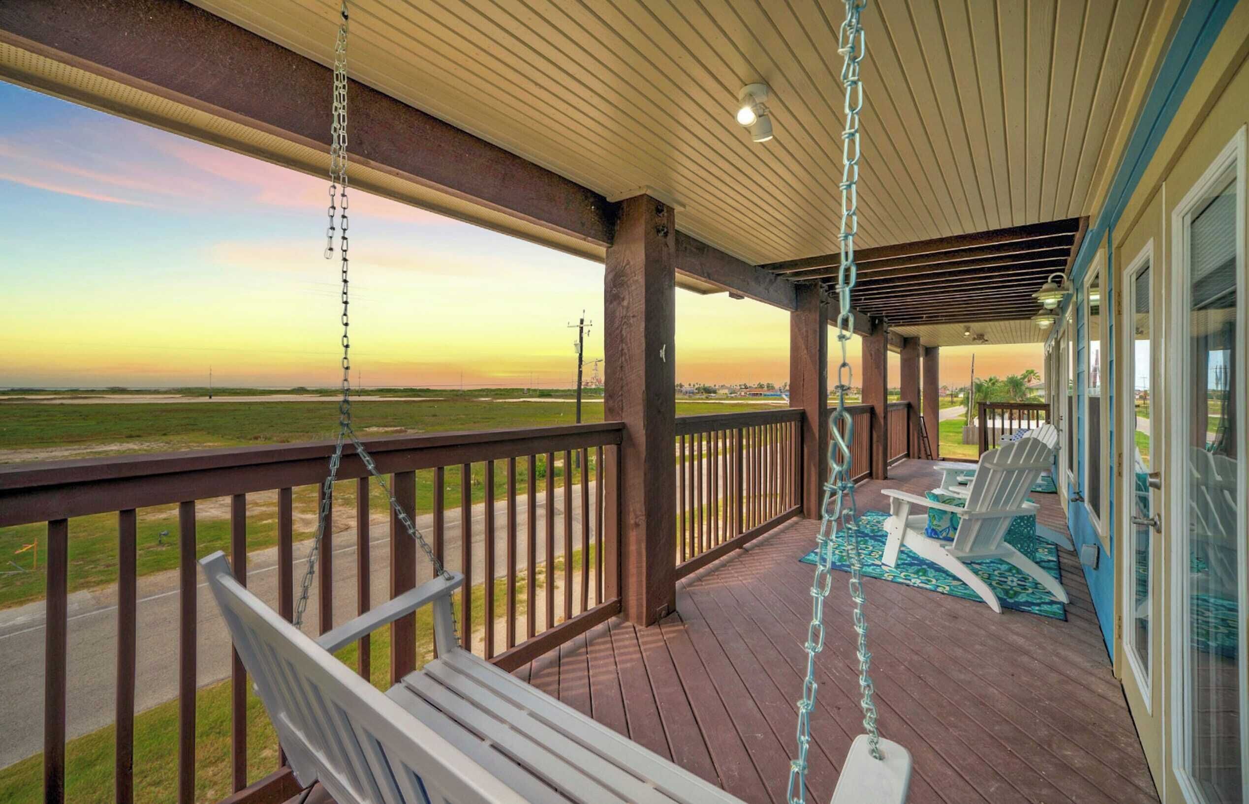 A covered porch with a swing chair and a lawn chair, overlooking a grassy field.