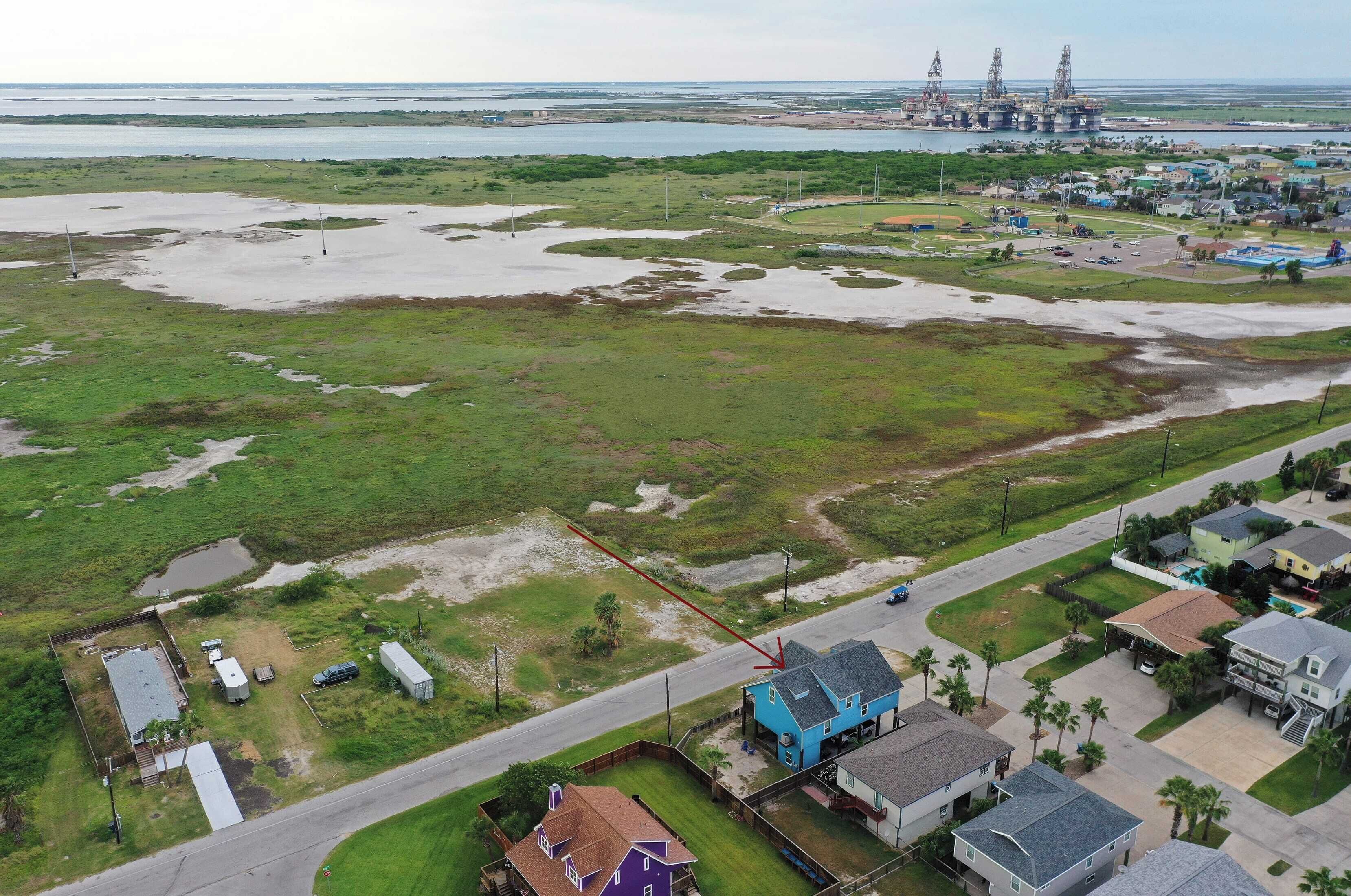 An aerial view of a residential area with houses, a road, and a large body of water with industrial structures in the background.