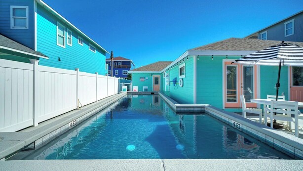 A swimming pool with a white fence, a turquoise building, and a small structure with a door and windows. - Peachy Palm by Avantstay Huge Private Pool! (Port Aransas)