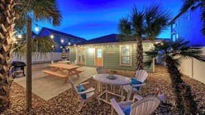 A patio area with a table and chairs, a wooden deck, and a building with a green exterior. - Peachy Palm by Avantstay Huge Private Pool! (Port Aransas)