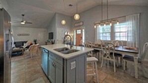 A kitchen with a central island, a dining area with a table and chairs, a ceiling fan, pendant lights, and a sign reading 'Beach House'. - Peachy Palm by Avantstay Huge Private Pool! (Port Aransas)