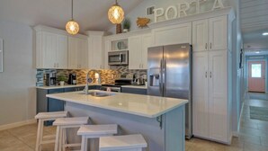 A modern kitchen with white cabinets, a stainless steel refrigerator, a microwave, and a large island with bar stools. - Peachy Palm by Avantstay Huge Private Pool! (Port Aransas)
