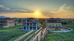 A coastal residential area with multi-story houses, a wooden boardwalk, and a sunset. - Beach Access Private Pool, Ping-pong, Firepit, Swings, & Views Tipsea Turtle (Port Aransas)