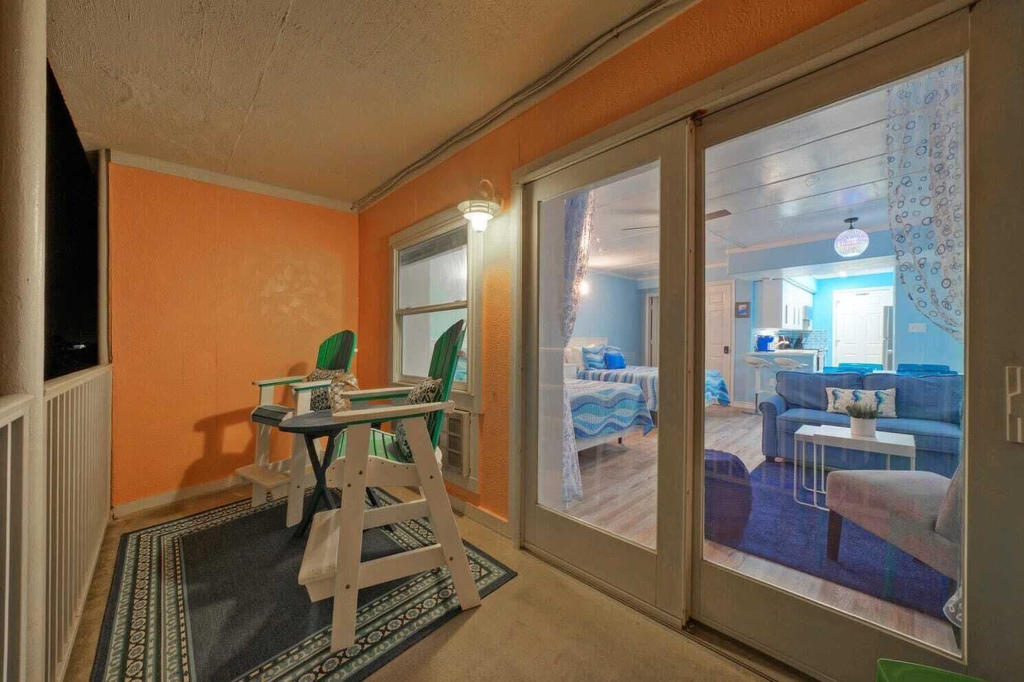 A room with a bed, a desk, and a chair. There is a sliding glass door leading to another room with a sofa and a table.