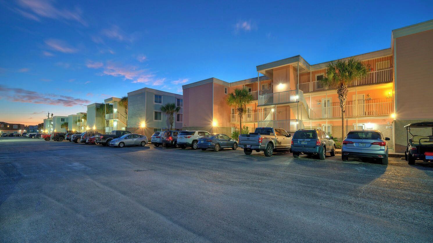 A parking lot with multiple cars and a row of buildings with balconies and palm trees.