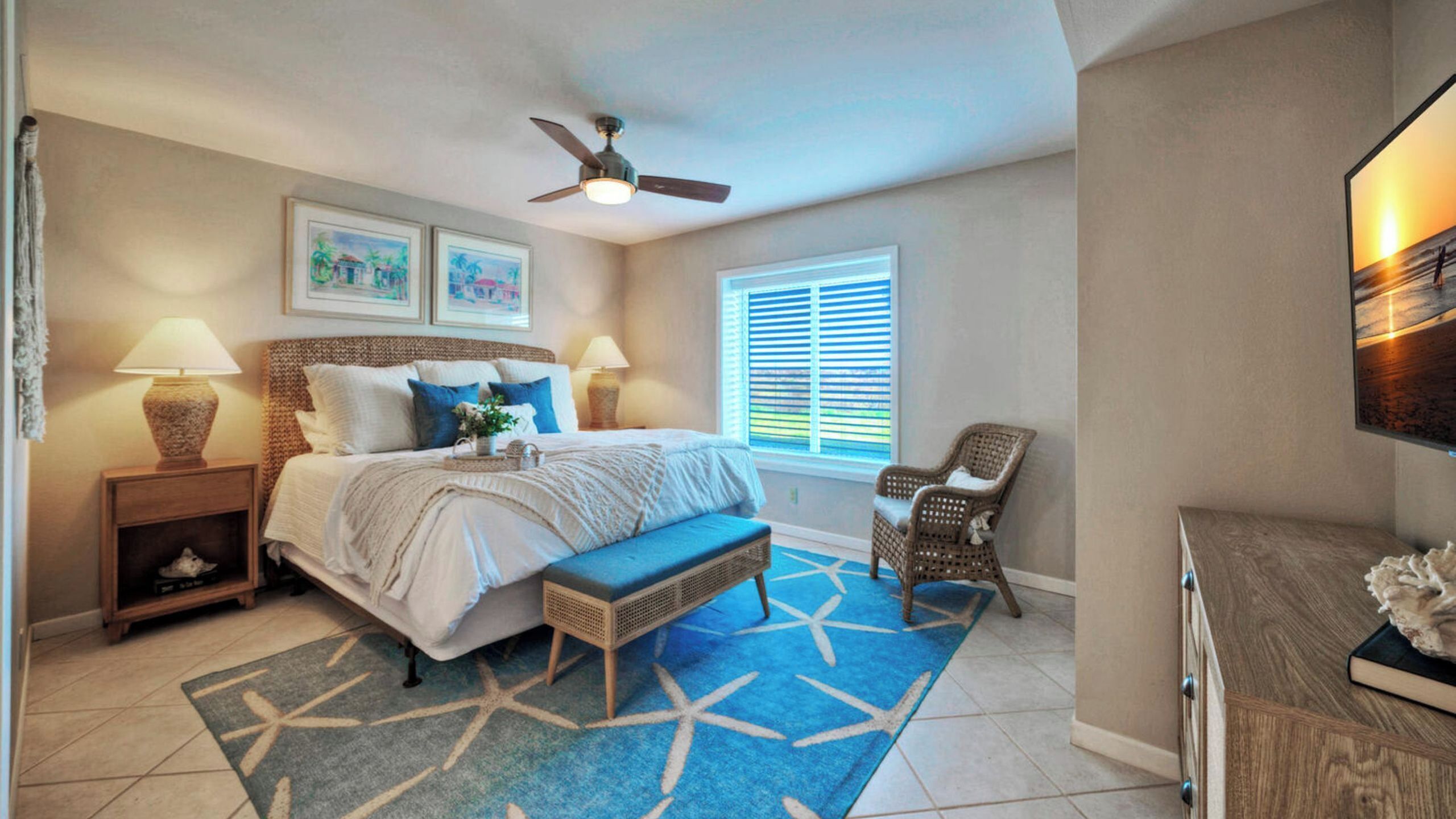 A bedroom with a bed, two bedside lamps, a ceiling fan, a window with blinds, a chair, and a TV on a wooden cabinet.