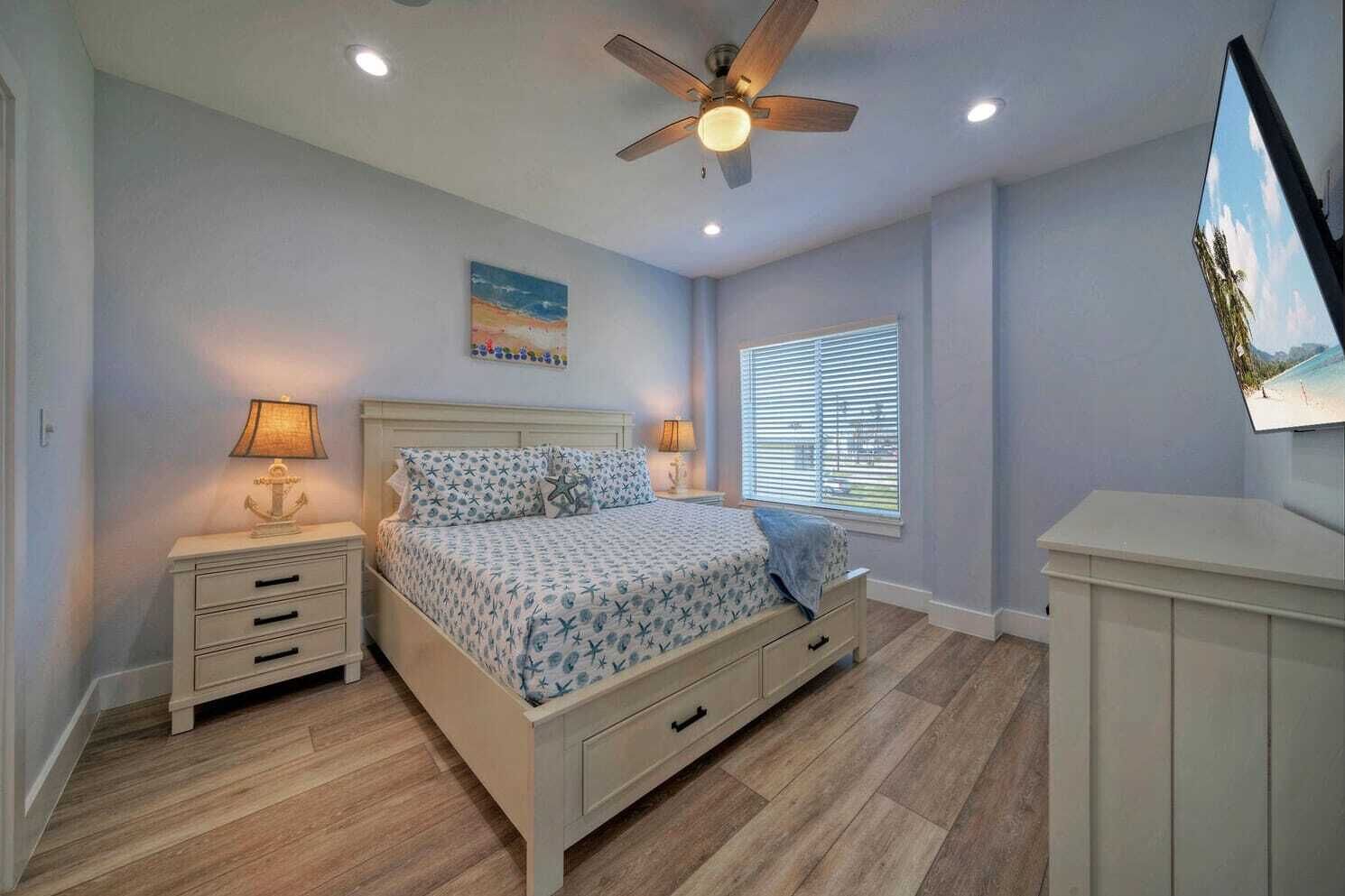 A bedroom with a bed, nightstands, a ceiling fan, a window with blinds, and a TV on the wall.
