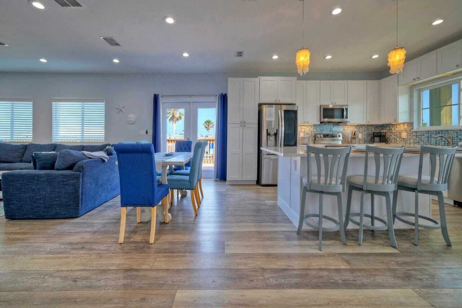 A modern living room with a blue sofa and dining area, a kitchen with white cabinets and a large island, and a view of the outdoors through the windows.