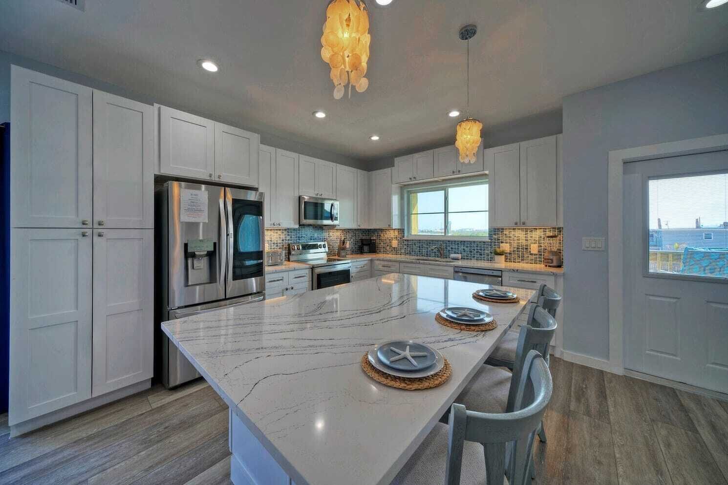 Una cocina moderna con armarios blancos, una isla grande de mármol, electrodomésticos de acero inoxidable e iluminación colgante.