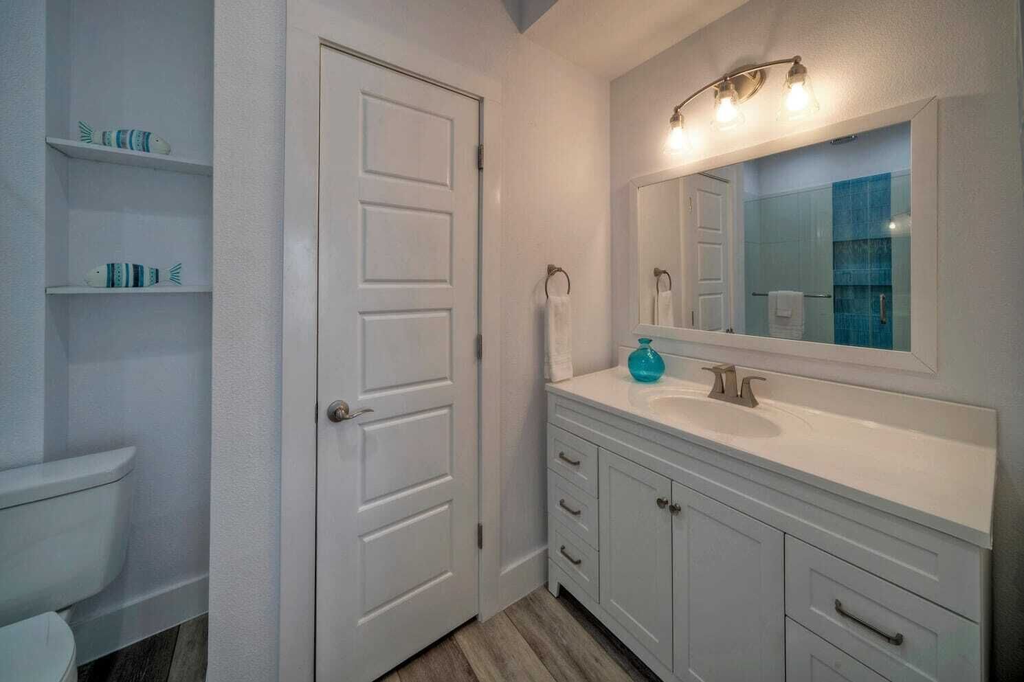 A bathroom with a white door, a white sink, a mirror, and a shelf with decorative items.