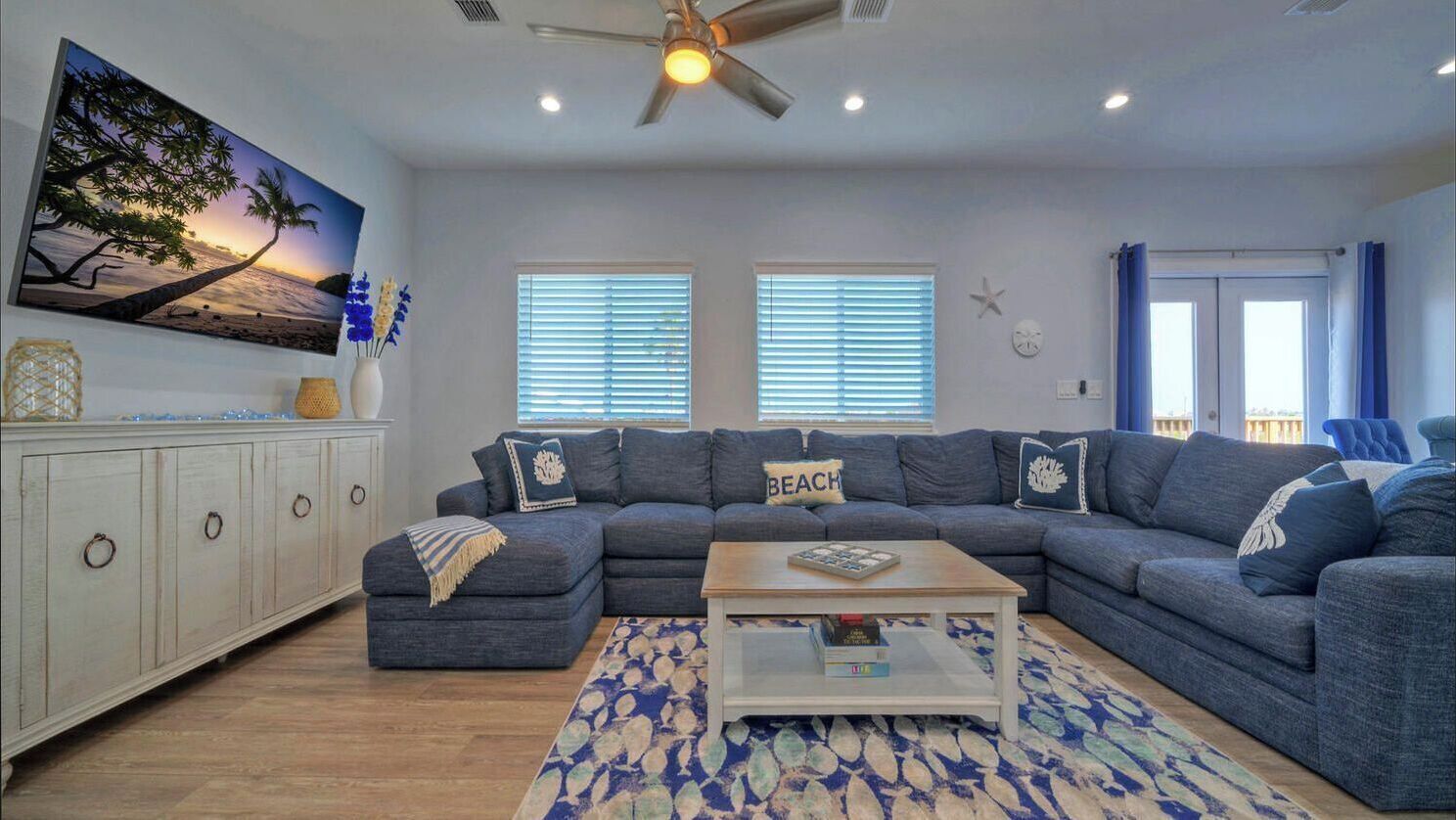 Ein Wohnzimmer mit einem Ecksofa, einem Couchtisch, einem Gemälde mit Strandmotiv und einem Deckenventilator.