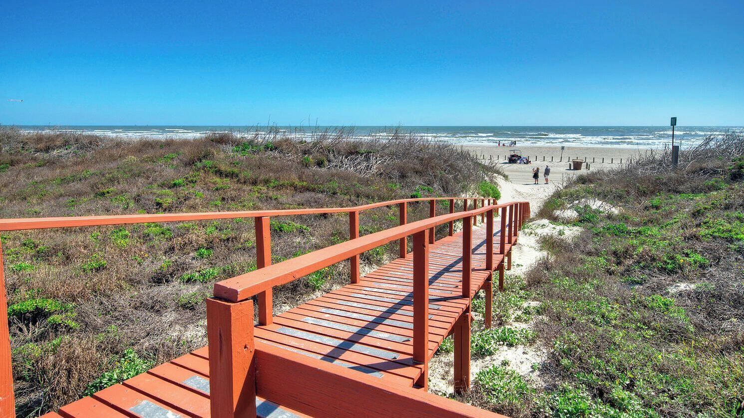 A wooden boardwalk leading to a beach with dunes and a clear sky.