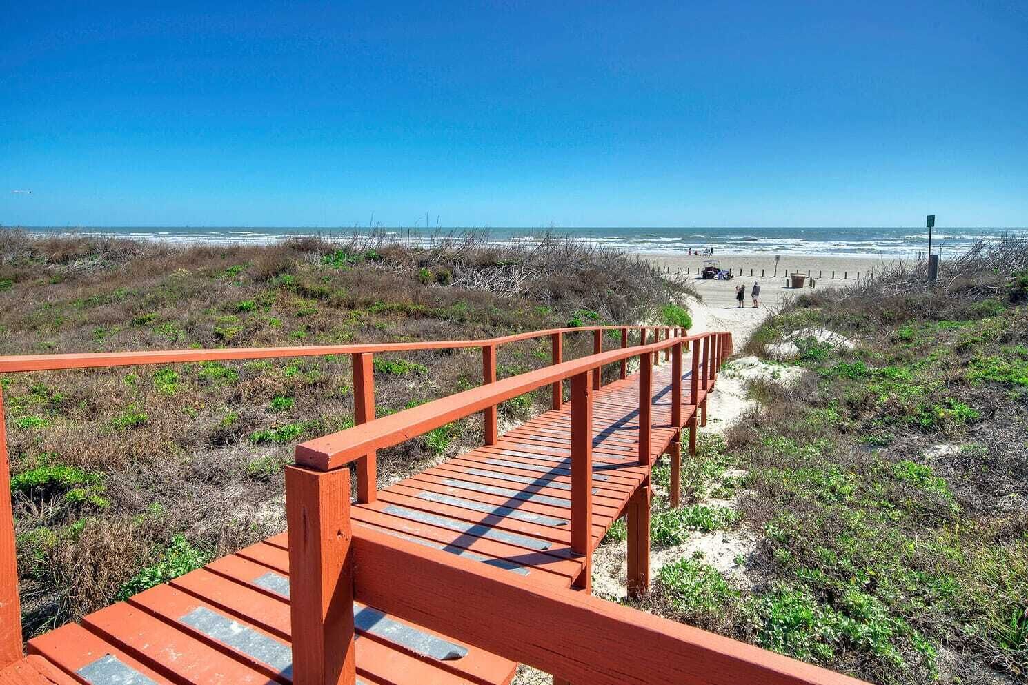 A wooden boardwalk leading to a beach with a clear view of the ocean and sandy dunes.