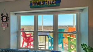 A room with a large window overlooking a beach, colorful chairs, and a sign reading 'BEACH HOUSE'. - Beach Boardwalk, Gulf View, Pool, Tennis+pickleball Beach Please! by Avantstay (Port Aransas)