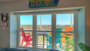 Ein Zimmer mit großem Fenster mit Blick auf einen Strand, farbenfrohe Stühle und ein Schild mit der Aufschrift 'BEACH HOUSE'.