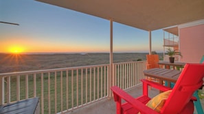 Ein Balkon mit Blick auf einen Sonnenuntergang über einer hügeligen Landschaft, ein Tisch mit Stühlen und ein Blumentopf.