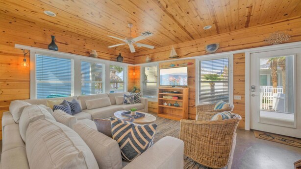 A cozy cabin interior with a sofa, wicker chair, ceiling fan, and large windows. - Gated Community w/ Pool 5mins to Beach Latitude Adjustment by Avantstay (Port Aransas)
