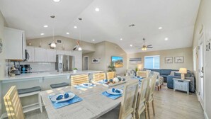 Cocina amplia con armarios blancos, una isla grande y electrodomésticos modernos. Hay una zona de comedor con una mesa larga y sillas, y una sala de estar con un sofá y una mesa de centro.