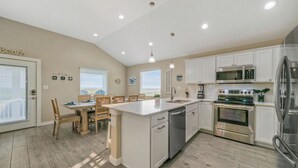 Una cocina con armarios blancos, una isla central, un comedor con sillas de madera y vistas a la playa.