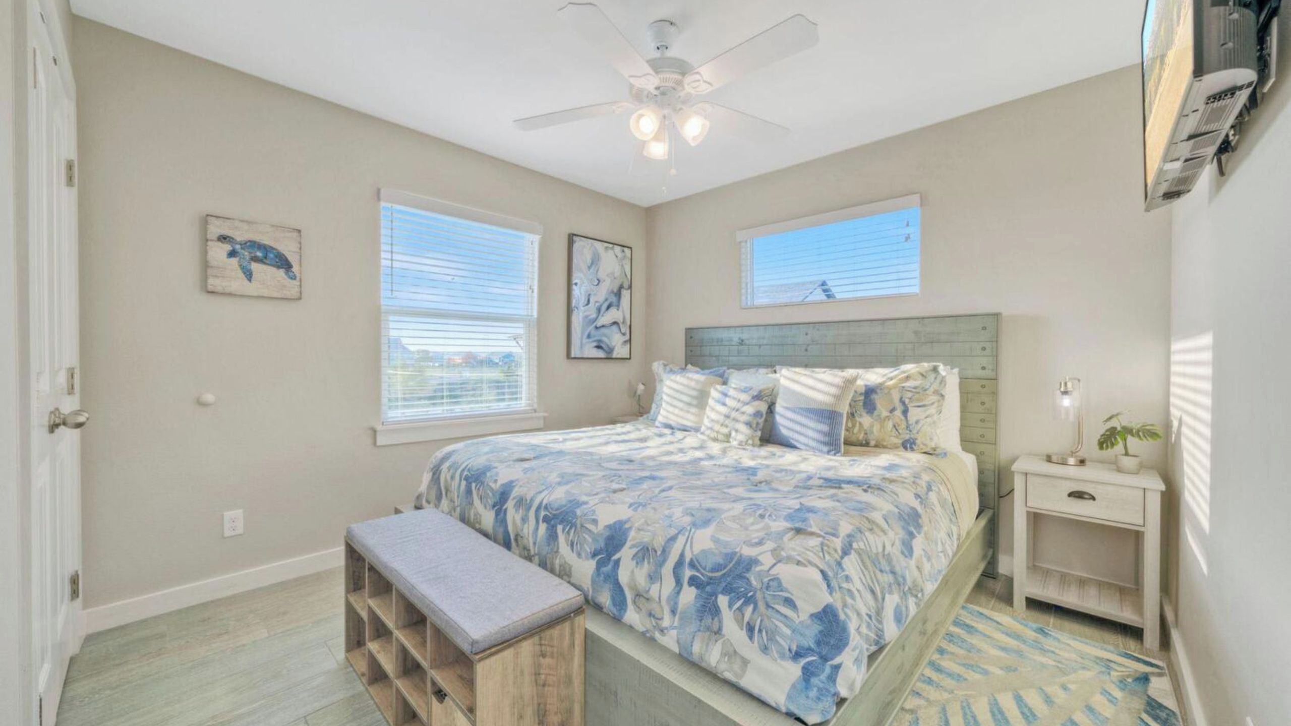 A bedroom with a bed, a bench, two windows, a ceiling fan, and a nightstand with a lamp.