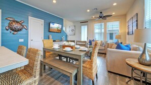 A dining area with a wooden table and wicker chairs, a living room with a beige sofa and blue cushions, and a ceiling fan. - Pool 1 Block to Alister + Old Town + Eateries Bluegrass Breeze by Avantstay (Port Aransas)