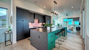 A modern kitchen with dark cabinets, a central island with bar stools, and a view to the living area. - Modern Design, 3 Mins to the Beach Boat Parking The Beach Box by Avantstay (Port Aransas)