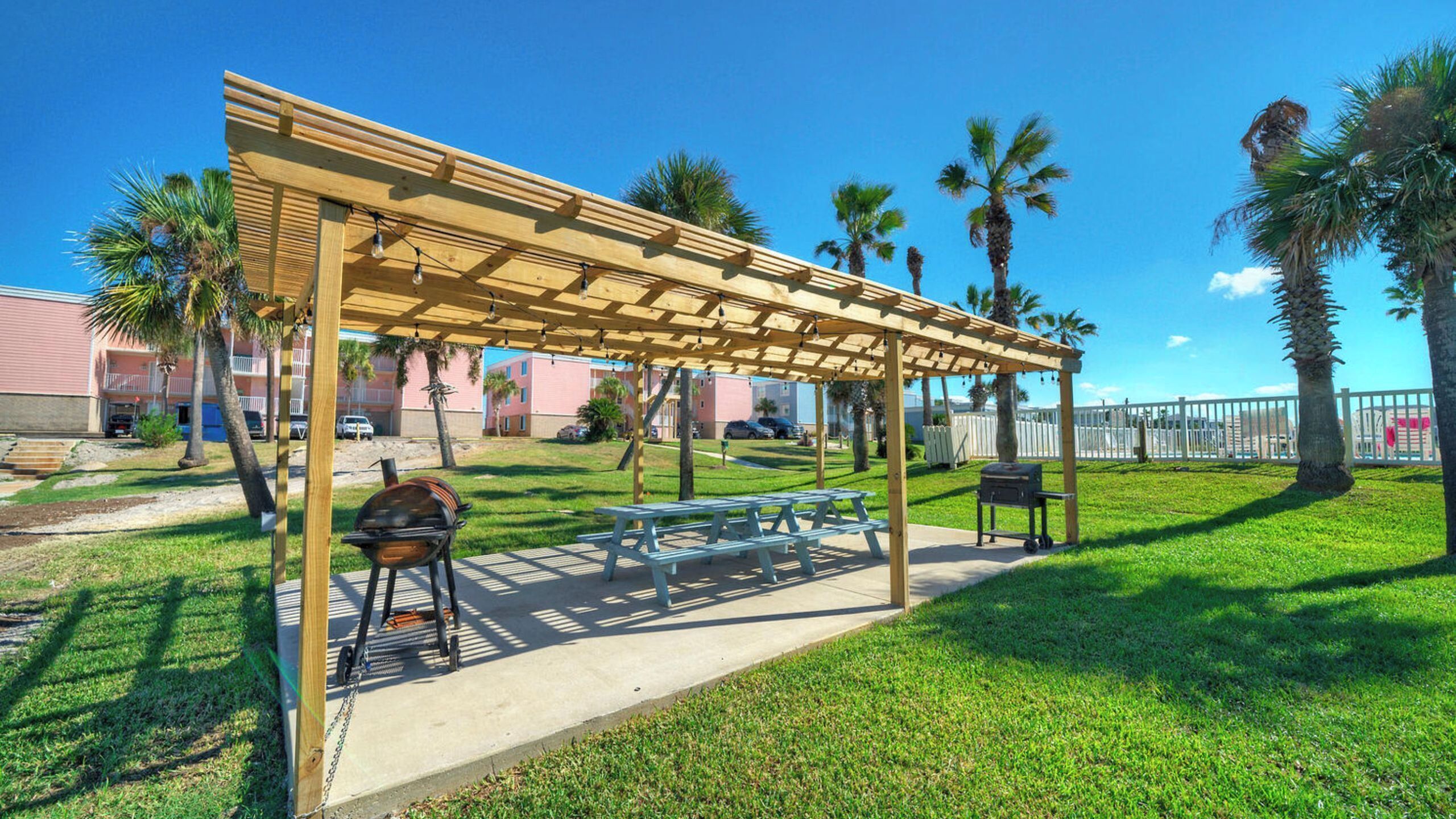 A covered outdoor area with picnic tables, a barbecue grill, and a grill cart, surrounded by grass and palm trees.