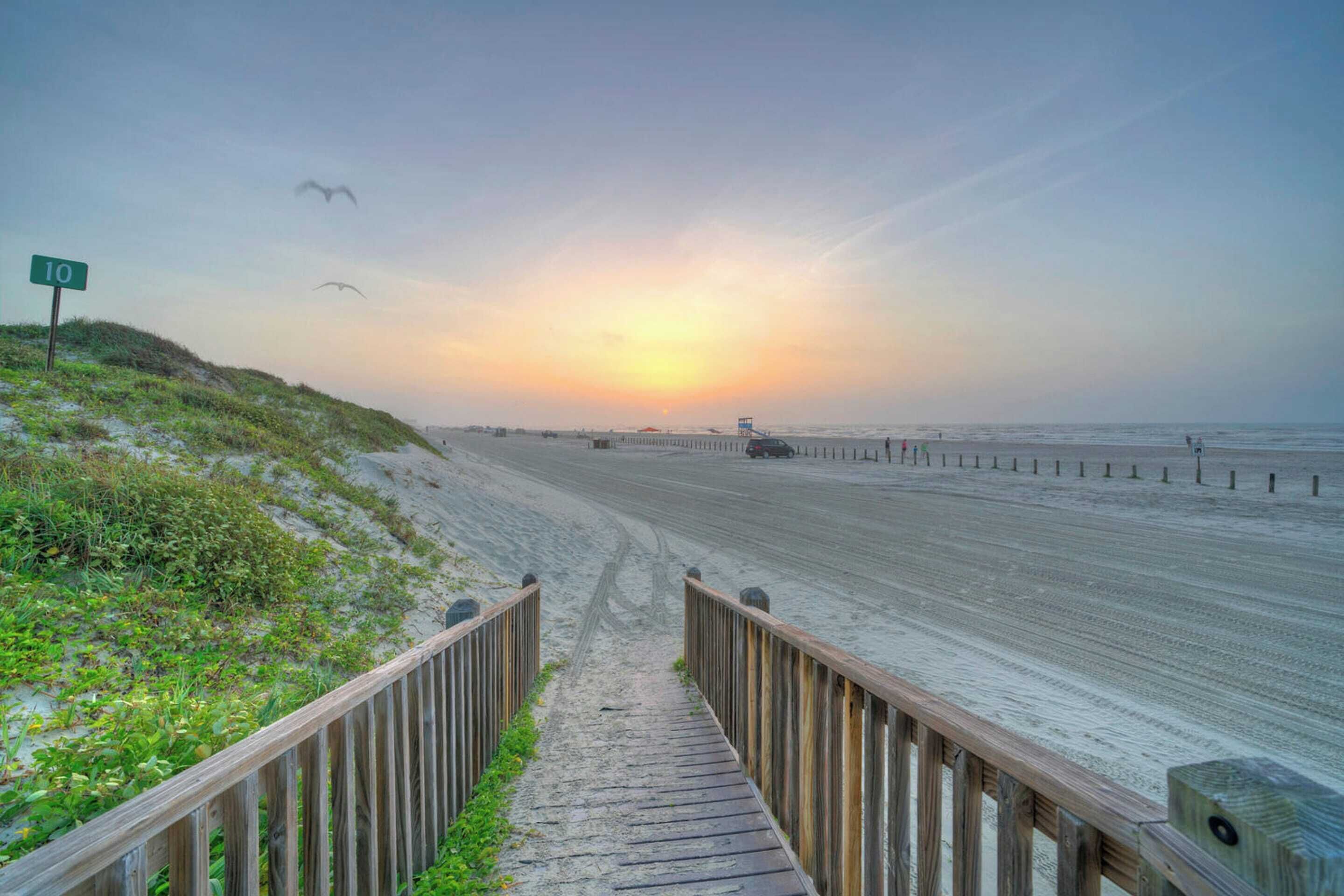 Une promenade en bois menant à une plage, avec un coucher de soleil en arrière-plan et une tour de surveillance des sauveteurs visible.