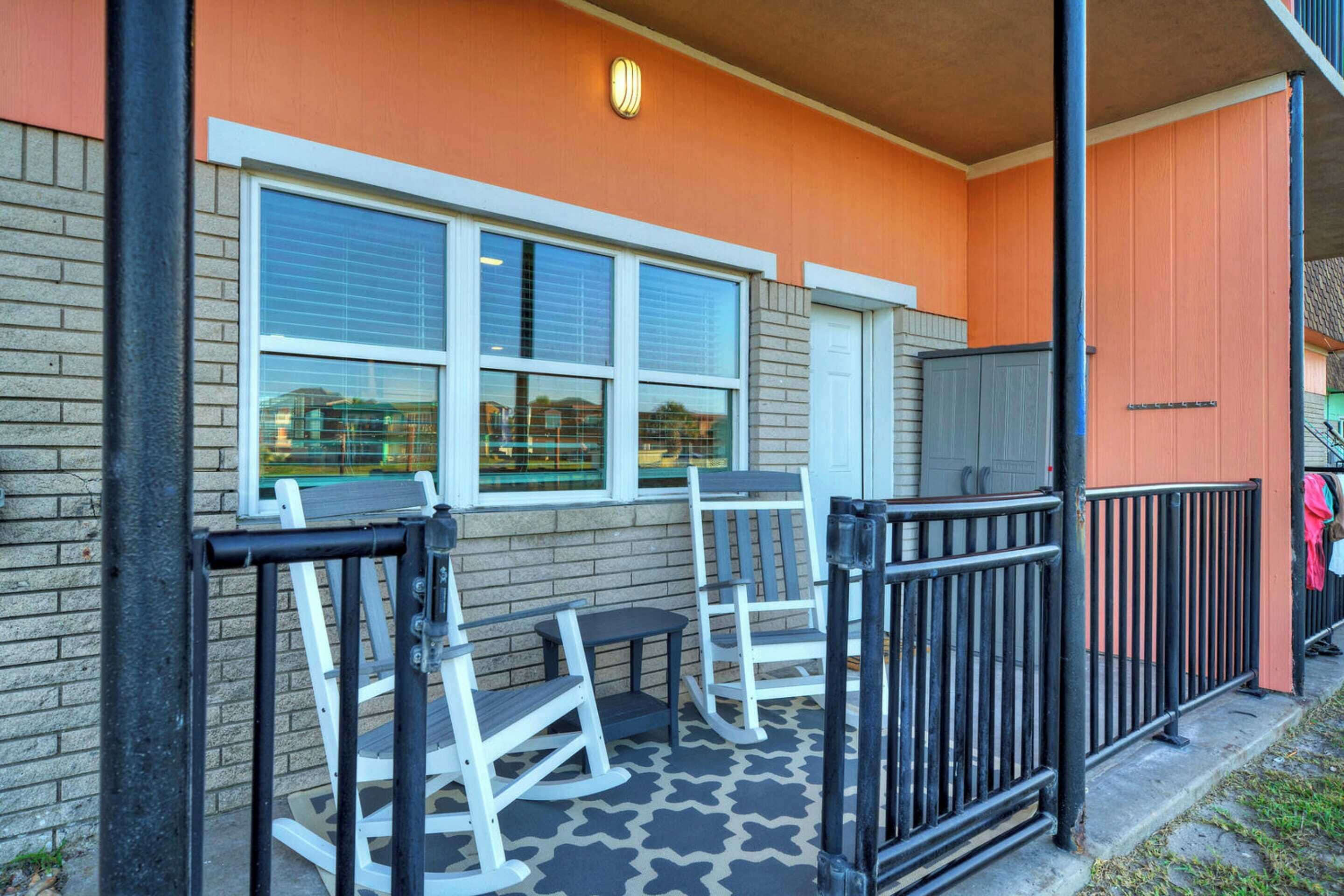 A building exterior with a brick facade, a window with blinds, a door, and a small outdoor seating area with chairs and a table.