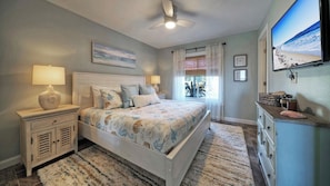 A bedroom with a bed, bedside tables, a ceiling fan, a window with curtains, and a wall-mounted TV. - Beachfront, Patio w/ Dune View, 2 Pools, Pickleball Cravin Waves by Avantstay (Port Aransas)