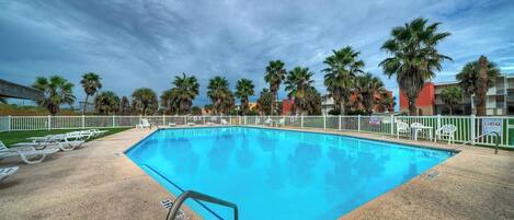 A swimming pool with a white fence, palm trees, and a cloudy sky.