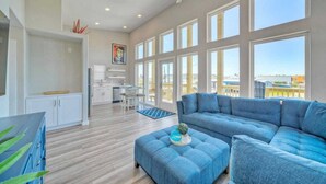A spacious living area with a large blue sectional sofa, a kitchen with white cabinets, and large windows offering a view of the outdoors. - Dean Marlin by Avantstay Stunning Old Town Home! (Port Aransas)
