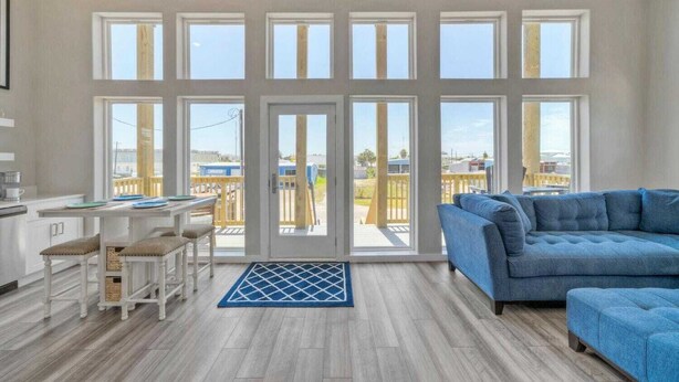 A spacious living area with a blue sofa, a dining table with chairs, and large windows offering a view of the outside. - Dean Marlin by Avantstay Stunning Old Town Home! (Port Aransas)