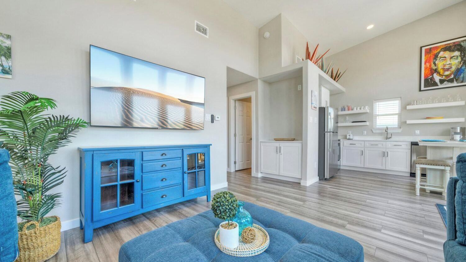 Una sala de estar moderna con un sofá azul, un cuadro abstracto grande y una cocina al fondo.