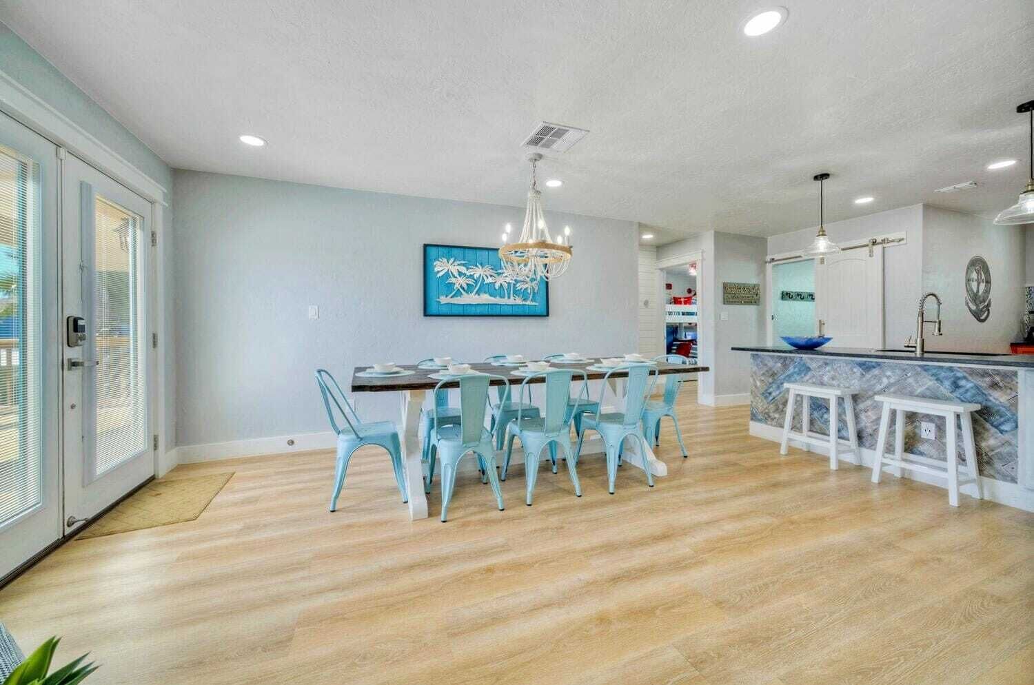 A spacious kitchen with a dining area, featuring a modern chandelier, a kitchen island with stools, and a large window with blinds.