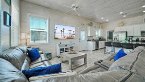 A spacious living room with a grey leather sofa, a coffee table, and a TV on the wall. There is a kitchen area with white cabinets and a dining space with a bar. - 2 Blocks to the Beach, Pool, Patio w/ Bar Awave From it All by Avantstay (Port Aransas)