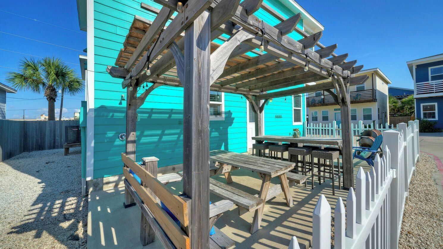 Une structure de pergola en bois avec une table et des bancs, un bâtiment turquoise et une clôture blanche à lattes.