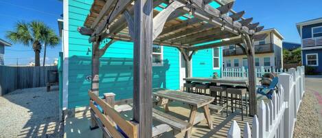 Une structure de pergola en bois avec une table et des bancs, un bâtiment turquoise et une clôture blanche à lattes.