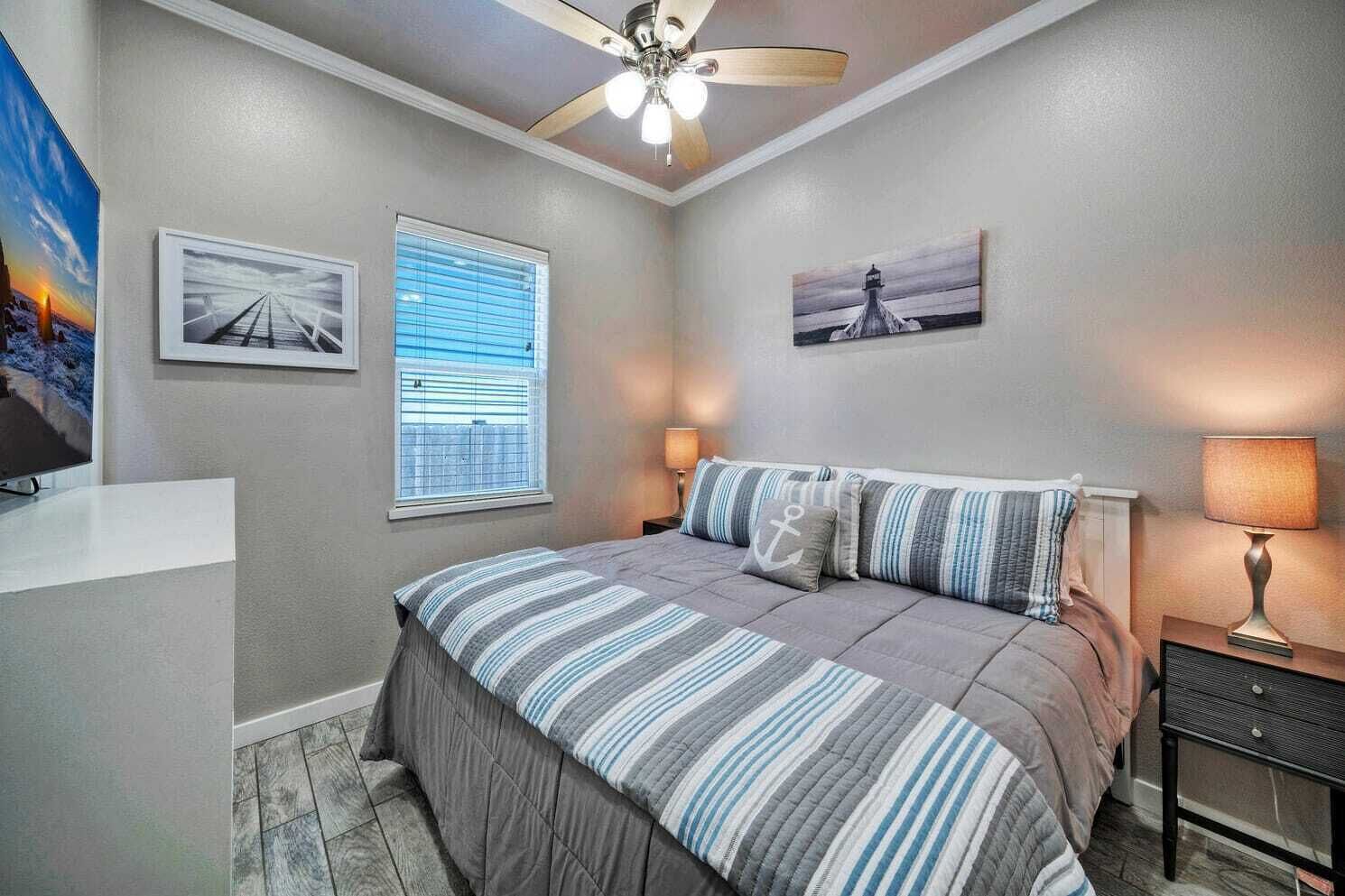 A bedroom with a bed, a ceiling fan, a window with blinds, and artwork on the walls.